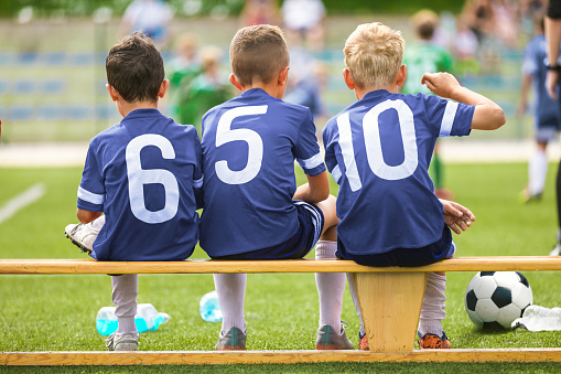 Football players in youth team sitting on wooden bench. Kids in school sports team. Boys in blue jersey shirts watching tournament game