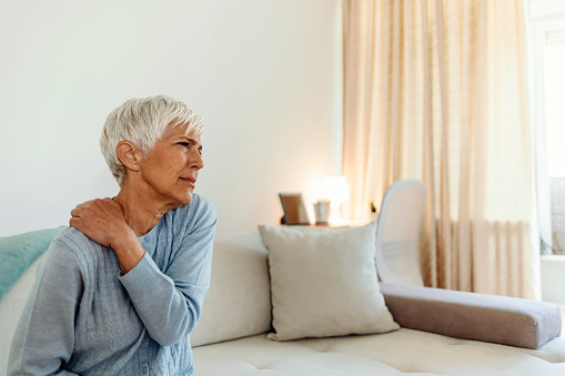 Stressed mature woman massaging her neck. Mature woman suffering from neck pain at home. Portrait of elderly woman having a neck pain in the living room. Stress and depression.