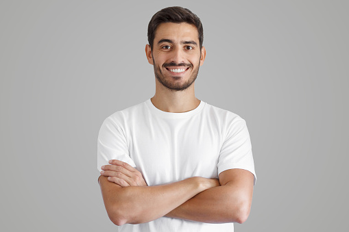 Portrait of smiling handsome man in white t-shirt, standing with crossed arms isolated on gray background