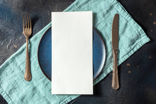 Dinner menu or invitation concept. A piece of white paper on a set table Dinner menu or invitation concept. A piece of white paper on a set table, with a fork, a knife, and a blue plate, overhead flat lay shot on black restaurant table card stock pictures, royalty-free photos & images