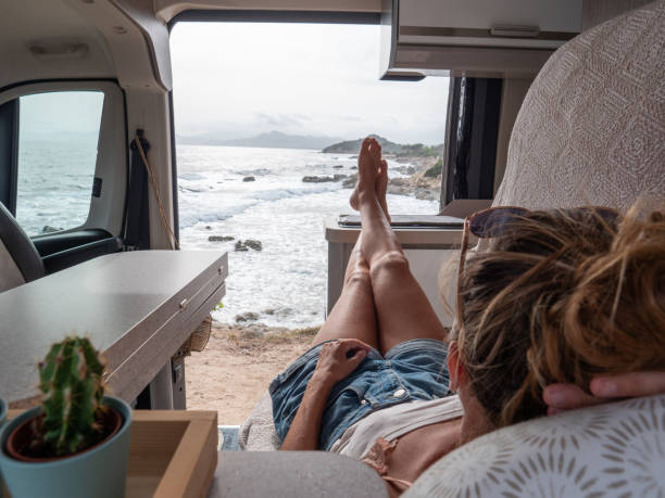 Woman in her camper van looking at ocean, she lies on the bench, side door open stock photo
