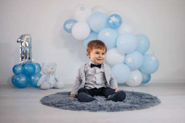 One year old child sitting on a rug next to balloons in celebration of his birthday.