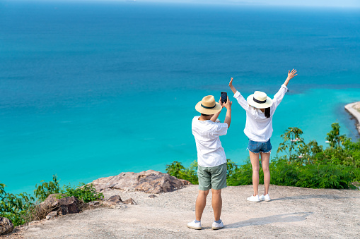 Asian family couple using mobile phone photography together while travel on tropical island mountain in summer sunny day. Husband and wife enjoy outdoor lifestyle on holiday travel beach vacation