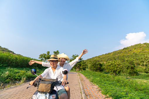 Happy Asian family couple riding motorcycle together while travel on tropical island mountain road in summer sunny day. Husband and wife enjoy outdoor lifestyle on holiday travel beach vacation