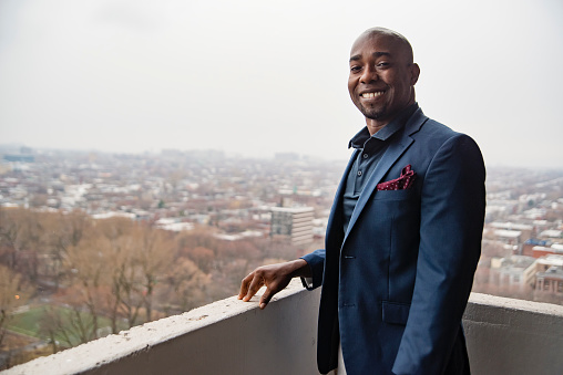 Portrait of mature black man on city balcony. He is wearing a blue polo and jacket and is looking at the camera with a smile. Foggy city in the background at springtime. Horizontal waist up outdoors shot with copy space.