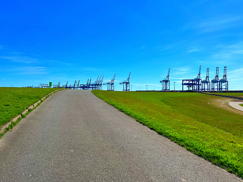 A path on the coast with cranes for containers in the background.