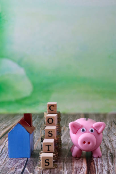 Stock photo showing a close-up view of piggy bank and blue cardboard house besides a staircase of stacked coins with wooden letter cubes spelling the word 'costs' on top coins. This is a family finances, real estate and holiday savings concept picture. imagen en primer plano de la escalera de monedas apiladas, cubos de letras de madera que deletrean la palabra 'costos' en la parte superior, además de la alcancía y la casa de cartón azul, enfoque en primer plano, fondo verde moteado, finanzas familiare