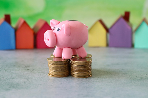 Stock photo showing a close-up view of piggy bank stood on piles of coins in front of a row of blue, orange, red, yellow purple and green cardboard houses. This is a family finances, real estate and holiday savings concept picture.