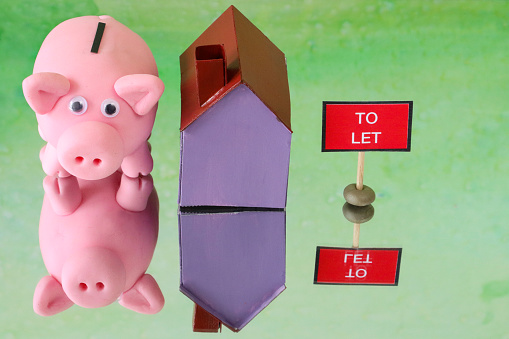 Stock photo showing a close-up view of a purple cardboard house besides piggy bank money box and a 'To Let' sign. This is a family finances, real estate and holiday savings concept picture.