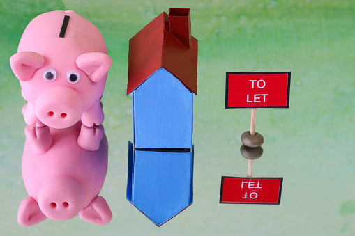 Stock photo showing a close-up view of a blue cardboard house besides piggy bank money box and a 'To Let' sign. This is a family finances, real estate and holiday savings concept picture.