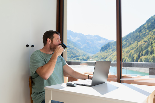 Man working from his apartment in the mountains. He is drinking coffee while is looking through the window during a work break. Working remotely.
