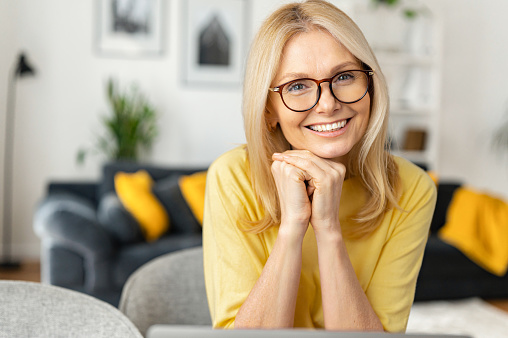 Charming middle-aged woman with long hair sitting in relaxed atmosphere. Mature 50s female resting at home, looking at camera and smiling with toothy smile