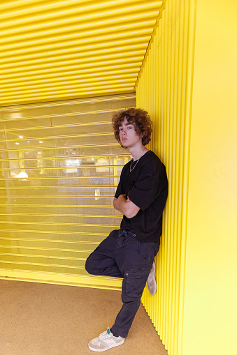 Vertical image of a young man leaning against a yellow wall with his arms crossed looking at the camera seriously.