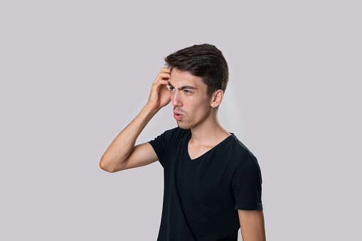 Portrait of a young man in a black T-shirt, side view, serious, scratching his head, indecisive ,confused. Studio shooting, gray background.