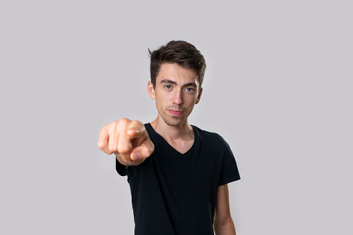 Portrait of a young man in a black T-shirt, front view, serious, index finger, pointing at you. Studio shooting, gray background.