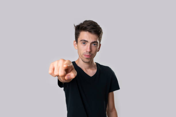 Portrait of a young man in a black T-shirt, front view, serious, index finger, pointing at you. Studio shooting, gray background. stock photo
