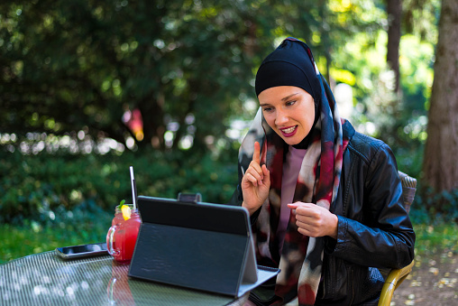 A Muslim woman in a hijab using a tablet in a street cafe next to the park.