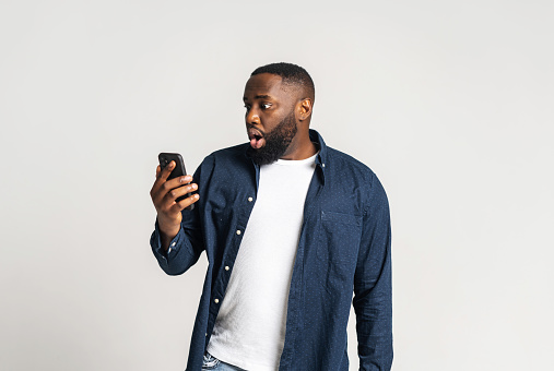 Shocked and dazed African-American guy holding smartphone and looks at screen, portrait of black pop-eyed man and open mouth watching with disbelieve at phone display isolated on grey