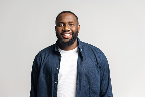 Smile. Cheerful glad African-American guy in casual blue shirt over white t-shirt looks at the camera isolated on white, happy black guy smiling with wide toothy smile. Studio shot