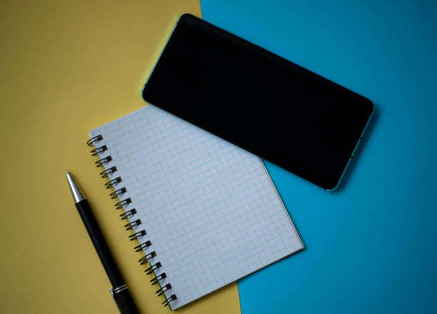 istock Office workplace minimal concept. Blank notebook with pencil, mobile phone on yellow and blue background. Top view with copy space, flat lay 1392577156