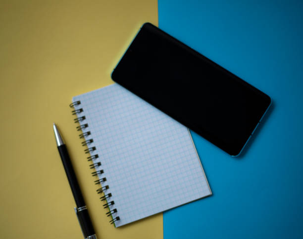istock Office workplace minimal concept. Blank notebook with pencil, mobile phone on yellow and blue background. Top view with copy space, flat lay 1392577157