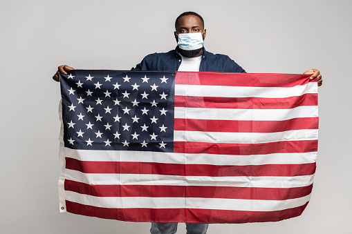 Serious African-American man in protective medical mask standing and looking at the camera, guy holding american flag spreaded in arms in front of him, isolated on gray background