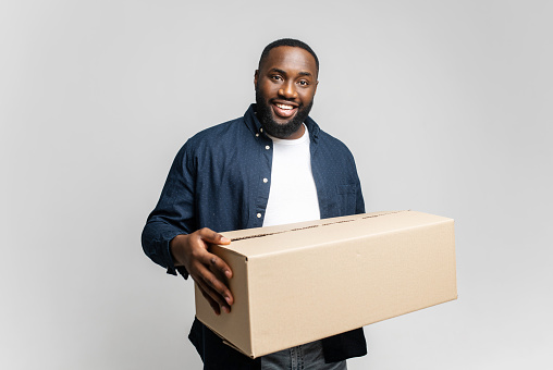 Glad young African-American man holding cardboard box, smiling and looking at camera isolated on gray background, delivery boy. Happy black guy moved in new house, relocation concept