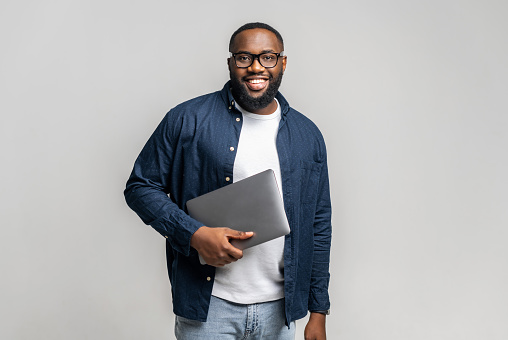 Smiling young African-American male entrepreneur stands in studio isolated on gray and carrying trendy laptop. Cheerful black guy in glasses and casual shirt looks at camera, holding closed laptop
