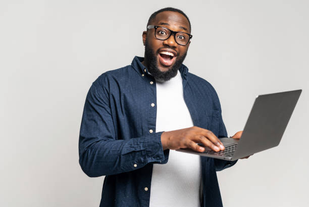 istock Black man standing with laptop on grey background, typing 1392578930