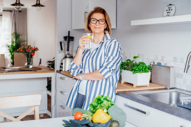 sana abitudine di bere acqua. sorridente donna di mezza età con un bicchiere di acqua pura con limone in piedi sulla sua cucina. controlla l'idratazione del corpo, traccia il bilancio idrico. vita sana. selectivbe focus. - foods and drinks simplicity purity clothing foto e immagini stock