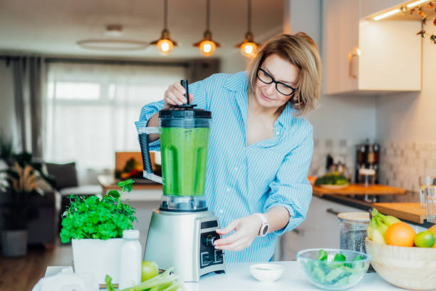 ブレンダーと緑の野菜を片手の中年女性が自宅でデトックスシェイクやグリーンスムージーを作っています。健康的なダイエット、食事、料理。自然なアンチエイジング、減量プログラム。� - smoothie drinking women drink ストックフォトと画像