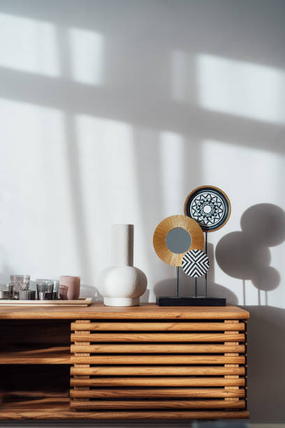 Vertical view of a modern minimalist Scandinavian style interior. Candles, ceramic vase and artistic home decor on a wooden console under sunlight and shadows on a white gray wall. Living room design
