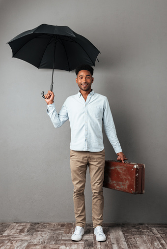 Full length portrait of a smiling young african man standing with open umbrella and suitcase isolated over gray background