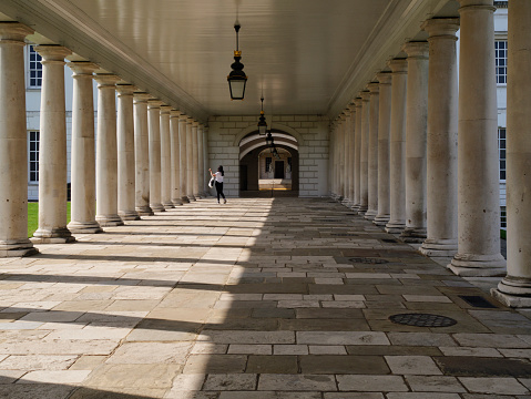 A woman waves to a friend from one of the dramatic colonnades at the National Maritime Museum in Greenwich, South East London, on a sunny spring day. The bright sun and the columns make interesting shadows.