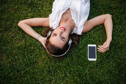 Picture of happy young woman lies on grass outdoors. Looking aside listening music.