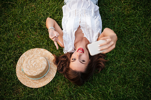 Image of cheerful young woman lies on grass outdoors. Looking aside make selfie showing peace gesture.