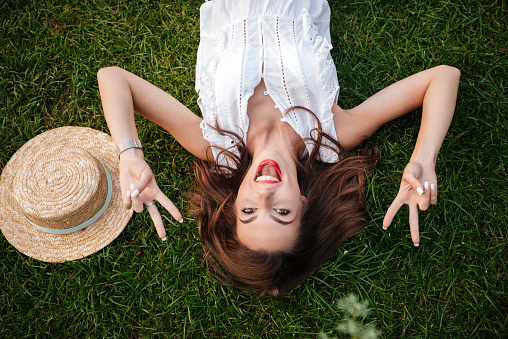 Image of cheerful young woman lies on grass outdoors. Looking camera showing peace gesture.