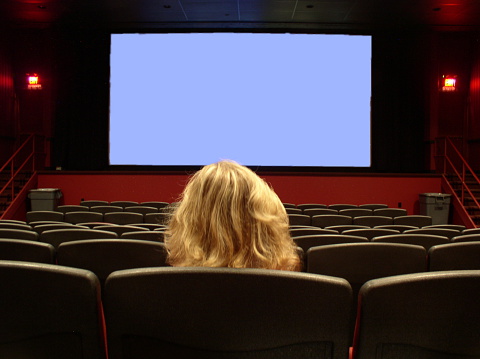 A girl sits alone in a movie theater waiting for the opening scene. Blue screen = your message. Focus is on the front seats with the girl slightly out of focus. Icon of lonelyness, doing things by yourself etc.