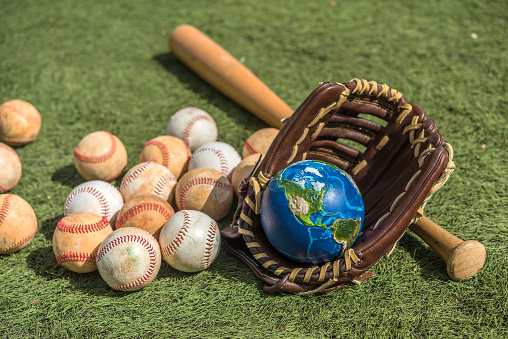 Planet Earth inside a baseball glove, wood bat, and balls on synthetic ground in a sunny morning. Conceptual photo on the globalization of baseball. \nNASA visual references (https://visibleearth.nasa.gov/images/74117/august-blue-marble-next-generation).