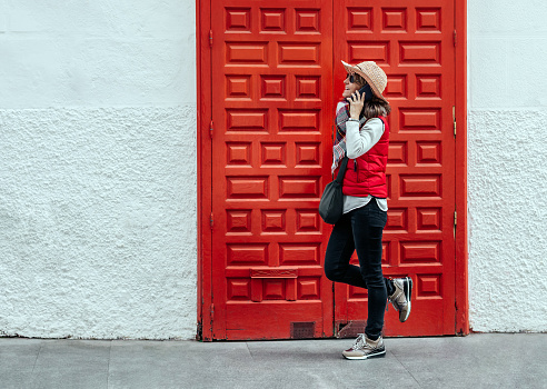 woman in hat and vest, talking on the telephone in front of a large red wooden door on a white background in madrid, spain. classic and modern concept. color contrasts.