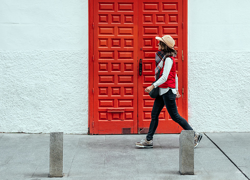 woman wearing a hat and vest walks in front of a large red wooden door on a white background in madrid, spain. classic and modern concept. color contrasts.