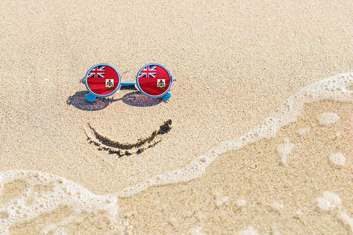 A painted smile on the sand and sunglasses with the flag of Bermuda. The concept of a positive and successful holiday in the resort of Bermuda.