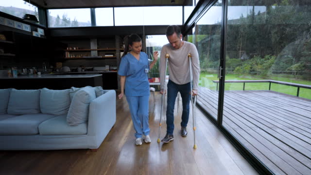 Female physiotherapist walking next to male patient using crutches at home