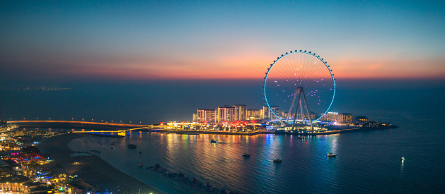 Panoramic view of Bluewaters island leisure spot in Dubai with large Ferris wheel seen from JBR beach in Dubai Marina area at blue hour. United Arab Emirates travel destination