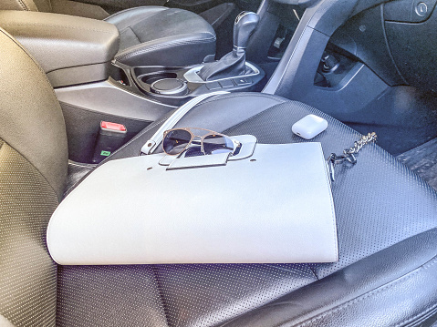 White women's beautiful fashionable leather bag and sunglasses with wireless headphones lie on the leather armchair of a good expensive car.