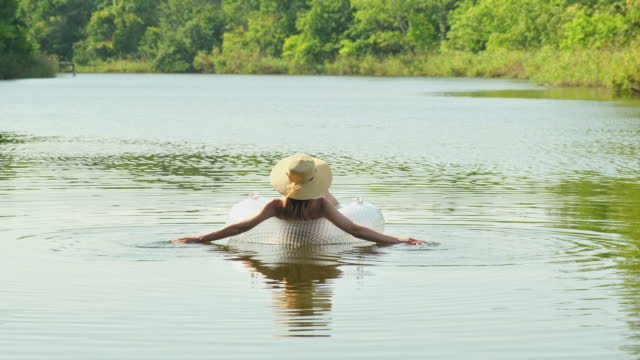 Woman in swimsuit and straw hat is floating with an inflatable circle in lake river pond. Girl rests on swimming ring in a pool with blue water. Concept of travel, vacation, weekends, holidays, relaxing on nature, rest, relaxation