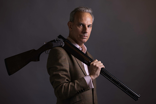 Suave, middle-aged man carrying a shotgun over his shoulder.