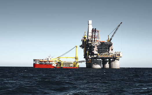 Oil rig offshore drilling platform and small support vessel. Blue clear sky, dark sea surface