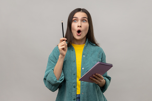 Excited woman writing creative idea into notebook, drawing sketch or taking notes in paper, raised arm, keeps mouth open, wearing casual style jacket. Indoor studio shot isolated on gray background.