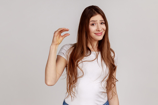 Portrait of unhappy woman with dark hair showing little size gesture with disappointed frustrated face expression, wearing white T-shirt. Indoor studio shot isolated on gray background.